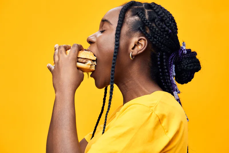 Young woman with braided hair enjoying a delicious burger against a vibrant yellow background, conveying joy and indulgence in fast food