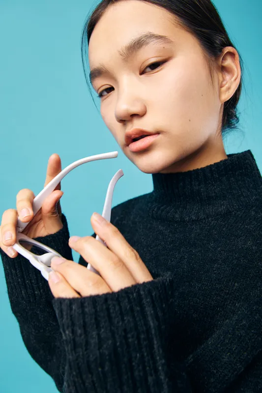 Young woman in black sweater holding white eyeglasses closeup with blue background. Portrait of thoughtful female model with clean skin and natural makeup in studio indoors.