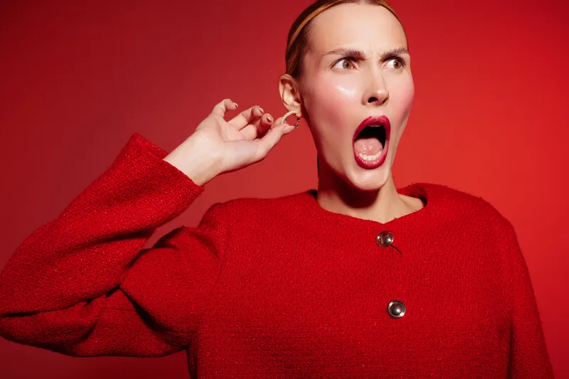 Surprised woman in red sweater with open mouth and hand near ear looks shocked on red background. Concept of surprise, emotion, reaction, and expression in fashion portrait.