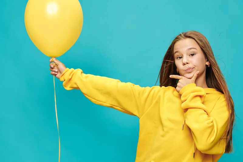 Girl in yellow hoodie holding yellow balloon on blue background posing with thoughtful expression for childhood, leisure and fun concept at studio.