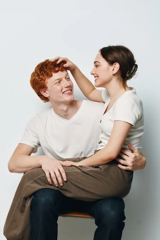 Young couple sitting together smiling and looking at each other with affection and happiness. Casual clothing and warm interaction in studio environment with light background.