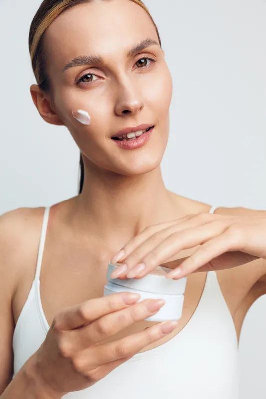 A young woman in a white tank top applies moisturizer to her cheek using her finger. The close-up portrait highlights skincare, beauty routine, and healthy glowing skin concept.