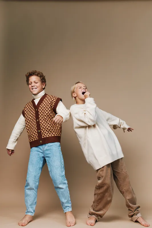 Two joyful children dance together in retro outfits, one wearing a patterned vest and jeans, the other in a cozy sweater and loose pants, against a warm beige studio backdrop.
