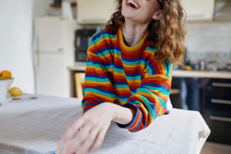 Cheerful woman indoors, laughing at a bright kitchen table, wearing a colorful striped sweater, warm natural light from a window, relaxed posture, soft breeze through a doorway, cozy home ambiance,