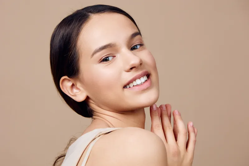 Smiling young model with a minimalist hairstyle, showcasing natural beauty and glowing skin against a soft beige background, highlighting a fresh and healthy lifestyle