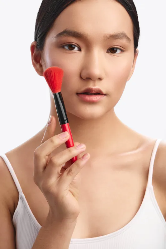 Asian woman applies makeup with a small brush in a clean studio, smiling softly as she focuses on grooming, creating a calm and polished beauty routine in a modern lifestyle setting.