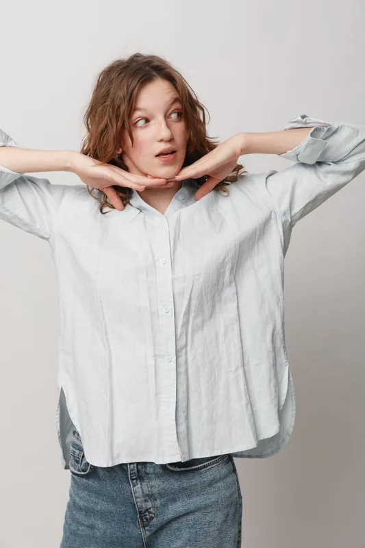 Young woman with curly hair wearing white shirt and jeans poses with hands near face looking sideways. Casual style, natural expression, isolated on light background.