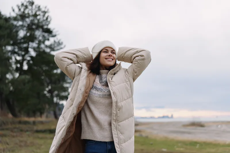 Woman outdoors wearing a long beige coat and beanie, smiling as she adjusts her puffer jacket in a serene landscape with trees, sand and a soft cloudy horizon.