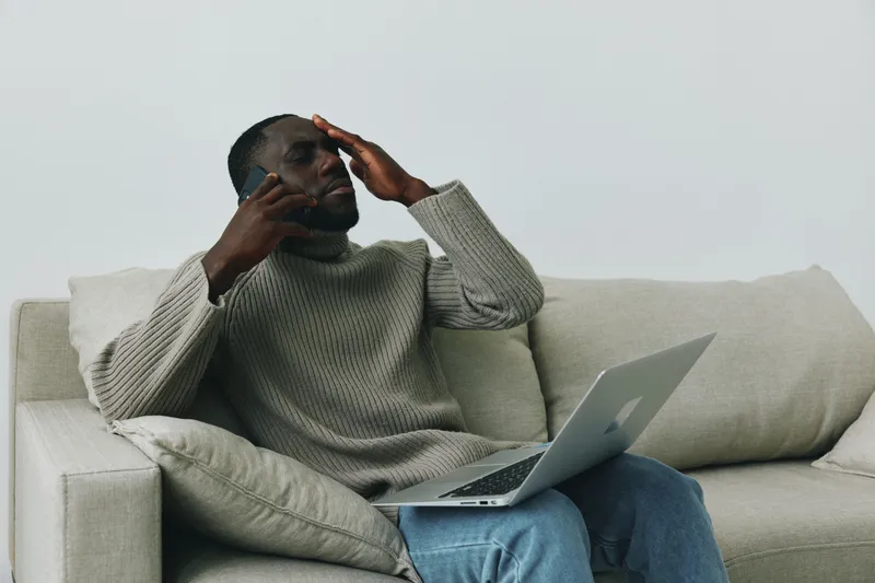 Young African man stressed while working on a laptop at home, wearing a cozy sweater, showcasing emotions of frustration and focus in a minimalistic setting