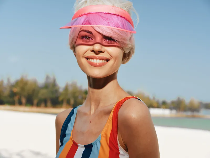 Bright portrait of a smiling woman on a sunny beach, wearing a colorful striped swimsuit and a pink visor, with clear blue sky and turquoise water in the background.