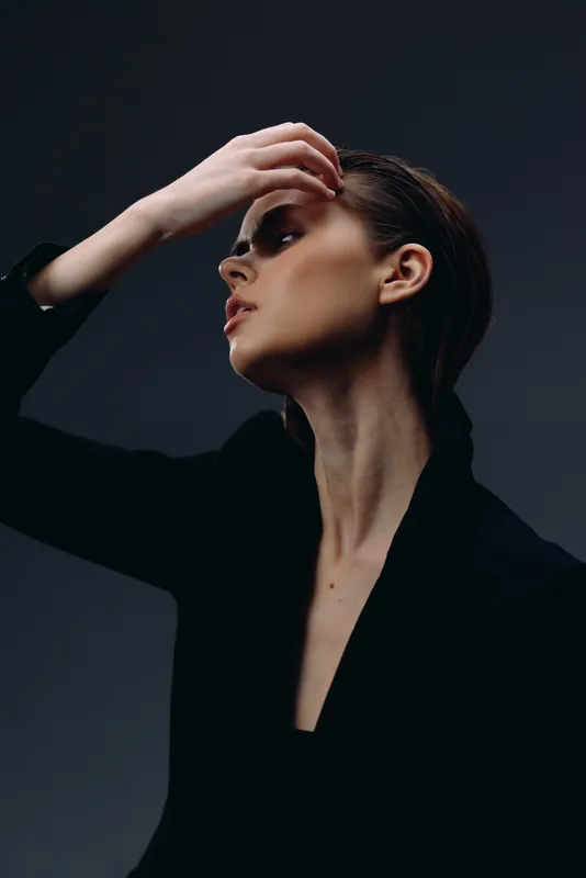 Elegant woman posing in dark clothing with dramatic lighting and contemplative expression against black background