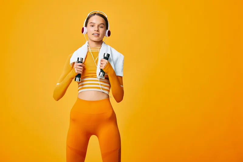 Fit young woman in orange workout set holds dumbbells with a towel over her shoulders and headphones on, posing confidently against a seamless bright orange studio background.