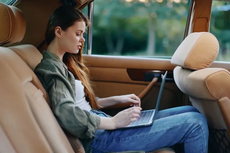 Young woman working on a laptop in the backseat of a car, showcasing a casual yet productive atmosphere with natural light and green background.