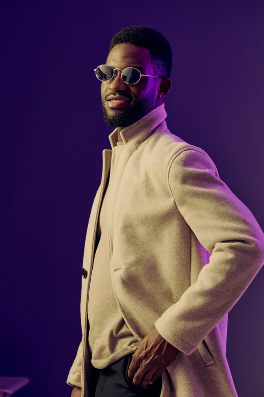 Stylish African man in sunglasses and a beige coat posing confidently against a purple background, exuding elegance and modern fashion aesthetics