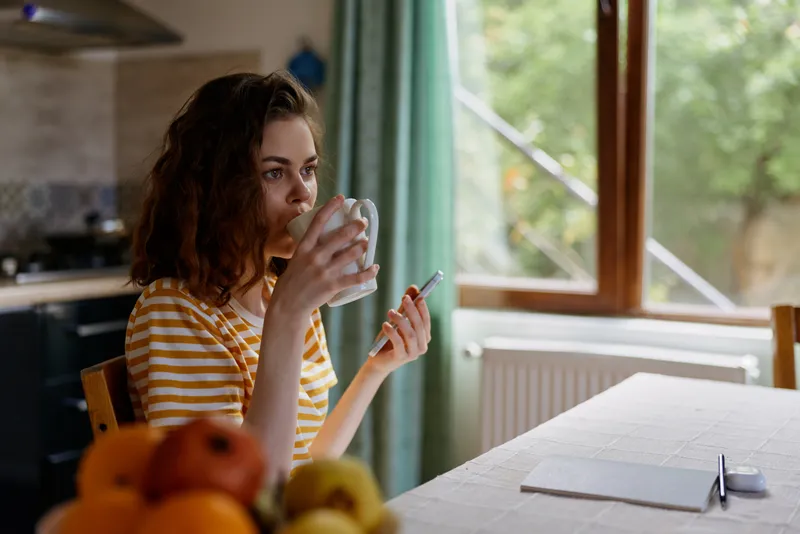 Calm woman in a striped shirt sits at a sunlit table, drinks coffee from a cup beside fruits and a notebook, capturing an intimate home lifestyle moment with warm, natural light.