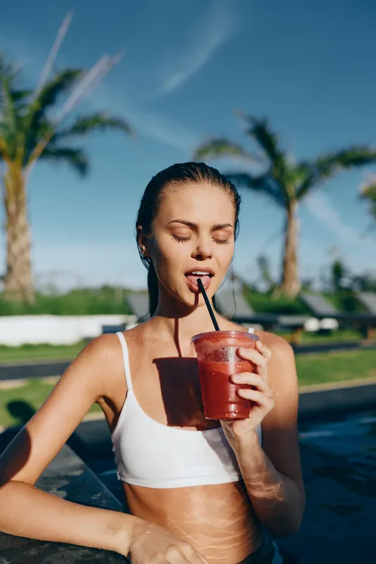 Young woman in a white bikini drinks a refreshing smoothie by the pool, relaxing on the edge under bright sun, palm trees in the background, a casual poolside lifestyle moment.