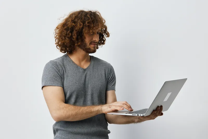 Young man with curly hair in a gray t shirt using a laptop, smiling, promoting remote work and digital lifestyle, minimalistic background