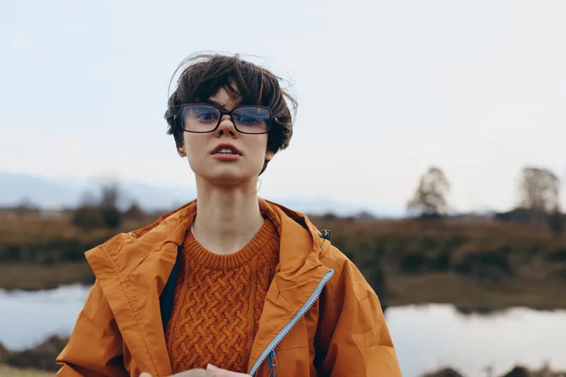Woman outdoors by river landscape, lifestyle portrait, orange jacket, wearing glasses, closeup face, calm expression, wind movement, nature background, casual mood, daylight
