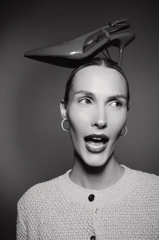 Young white woman with fair skin wearing textured jacket has high heel shoe on her head, looking sideways in studio.