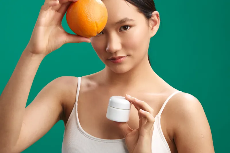 asian woman holds an orange near her head and applies cream, posing with a bright, fresh look in a minimal studio setup to convey wellness, beauty, and self care in a modern lifestyle scene.