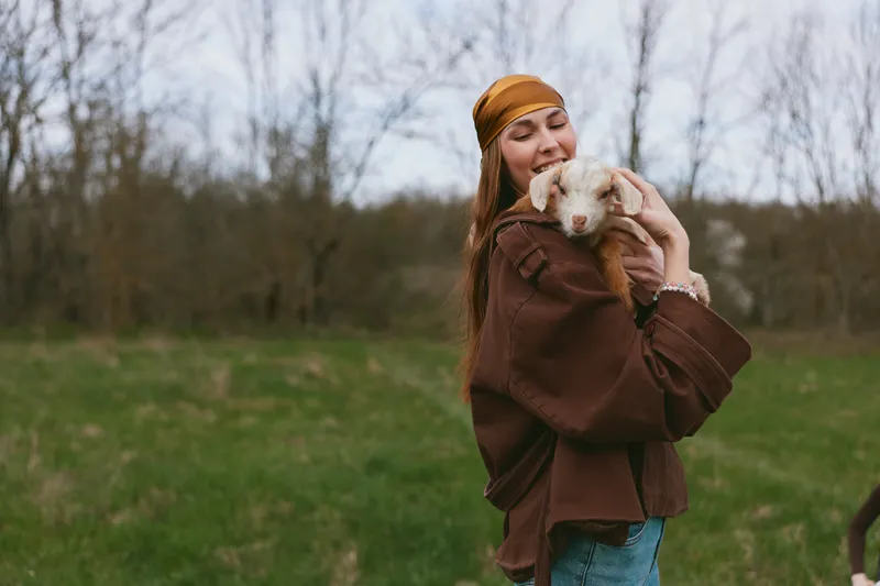 Smiling woman in a brown jacket outdoors gently holds a goat kid, capturing a warm rural lifestyle moment, connection and care in soft natural light for a tender everyday scene.