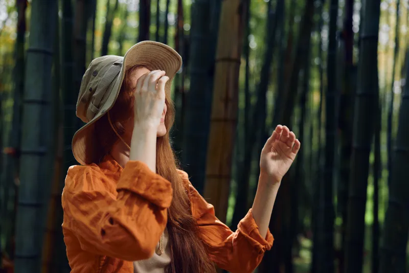 Woman in a straw hat stands in a bamboo forest, redhead portrait with orange blouse and natural light, touching hat with hand, serene outdoor nature scene and relaxed travel mood