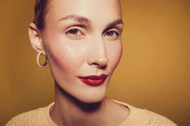 Close-up portrait of confident woman with red lipstick and hoop earrings on yellow background. Concept of beauty, fashion, makeup, and elegance with soft lighting and warm tones.