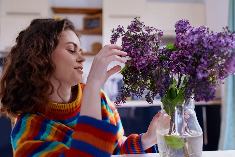 A young woman with curly hair in a colorful striped sweater arranges purple lilac flowers in a clear glass vase, creating a cozy home floral moment indoors while soft daylight streams through a