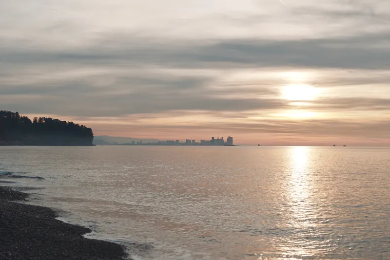 Sunset over calm water near a rocky shore with distant skyline on the horizon, soft light reflecting on the surface creating a tranquil coastal scene.