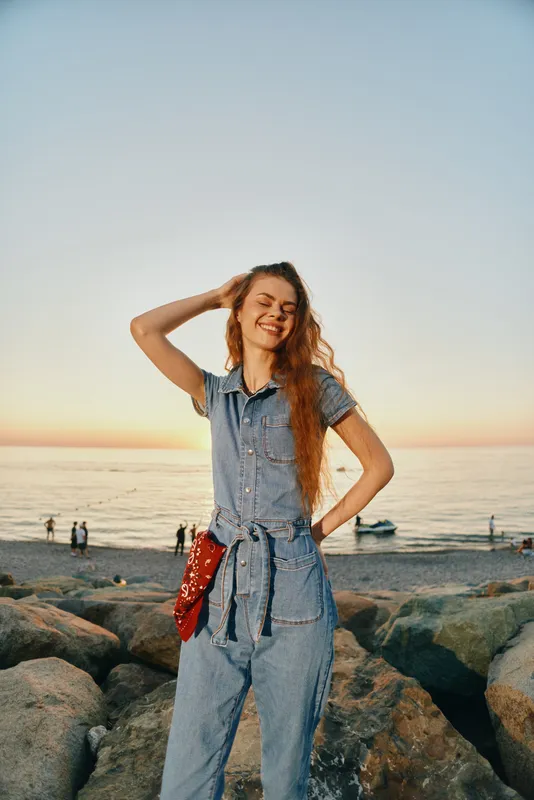 Lifestyle woman in modern boho-western denim street style stands smiling on rocky beach at sunset with film color tones, casual and relaxed summer mood.