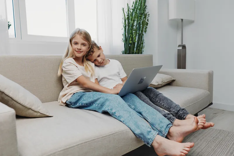 Two smiling children sitting barefoot on a sofa with a laptop, enjoying leisure time in a bright modern living room with natural light and houseplants.