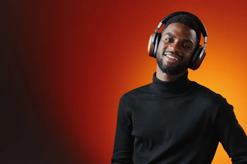 Stylish African man in headphones enjoys music, smiling against a vibrant orange background, representing joy and creativity in modern lifestyles