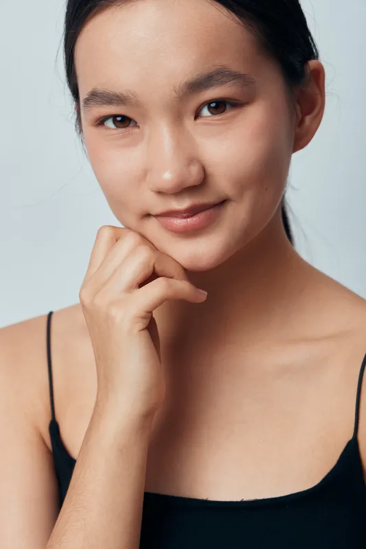 Young woman with natural makeup and clear skin posing with hand near chin against plain background. Beauty portrait of confident Asian female model wearing black top and soft smile in studio.