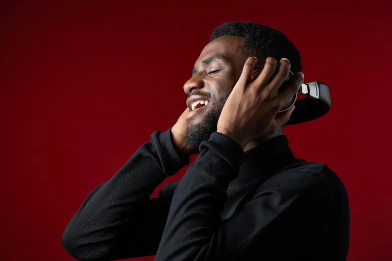 Smiling African man enjoying music with headphones against a red background, showcasing joy and relaxation in a stylish black outfit