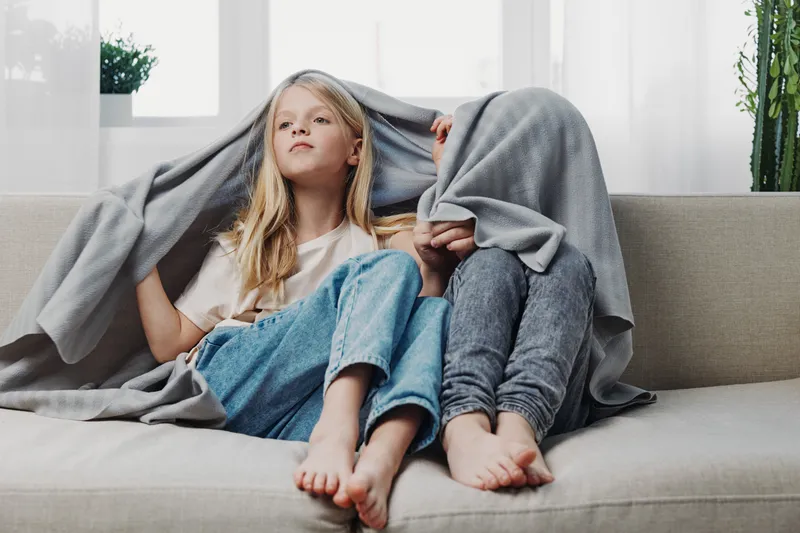 Two children sitting barefoot on a sofa covered with a gray blanket, playing and enjoying cozy time in a bright living room with natural light and plants in the background.