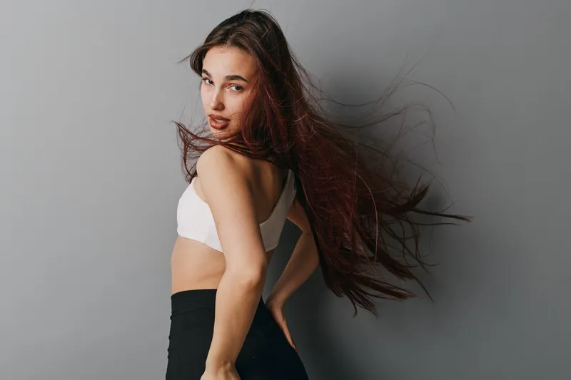 Young woman with long hair wearing sportswear posing against gray background. Fitness model in white top and black leggings with dynamic hair movement and confident expression.