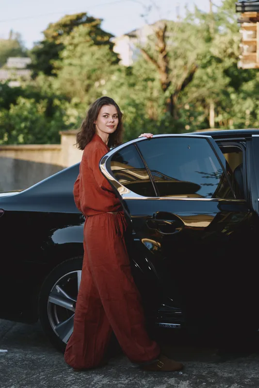 Woman in rust jumpsuit stands beside a sleek car, posing for a lifestyle shot, outdoor urban setting, confident expression, candid fashion moment, daylight, natural light, street style