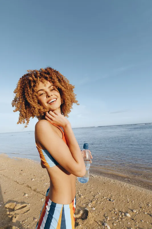 Woman on beach by ocean, smiling with curly hair, wearing striped swimsuit, sunny summer travel mood, relaxation pose with soft light, peaceful shoreline background, golden sand in distance