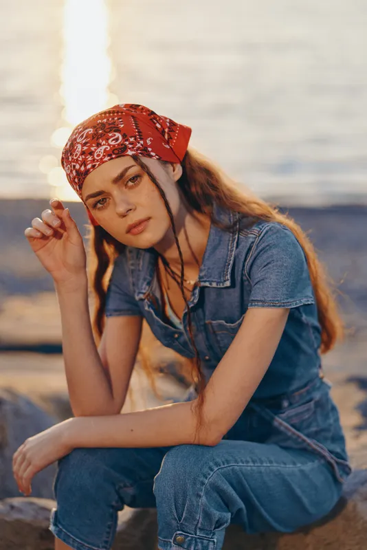 Lifestyle woman in modern boho-western denim street style with a red bandana, film color tone, relaxed pose by the beach at sunset, casual and stylish appearance.