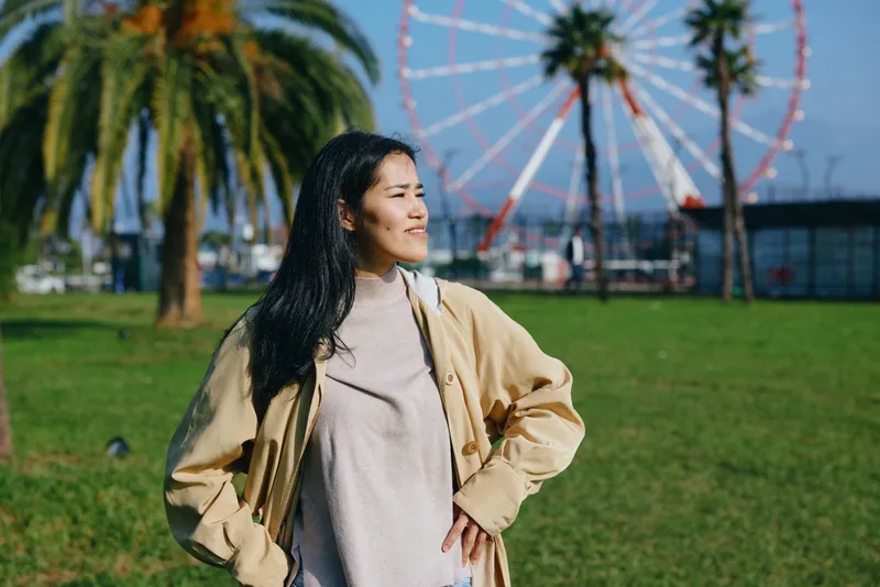 Woman in a casual jacket and sweater stands in a sunny park with a ferriswheel and palm trees in the background, confident and relaxed pose, candid outdoor scene.