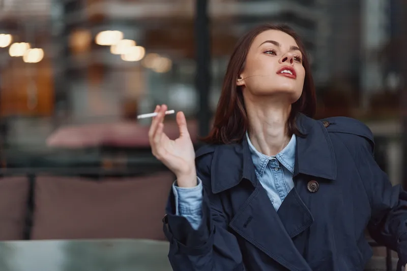Young woman with long brunette hair wearing a dark trench coat smokes a cigarette indoors with a confident and relaxed expression on her face.