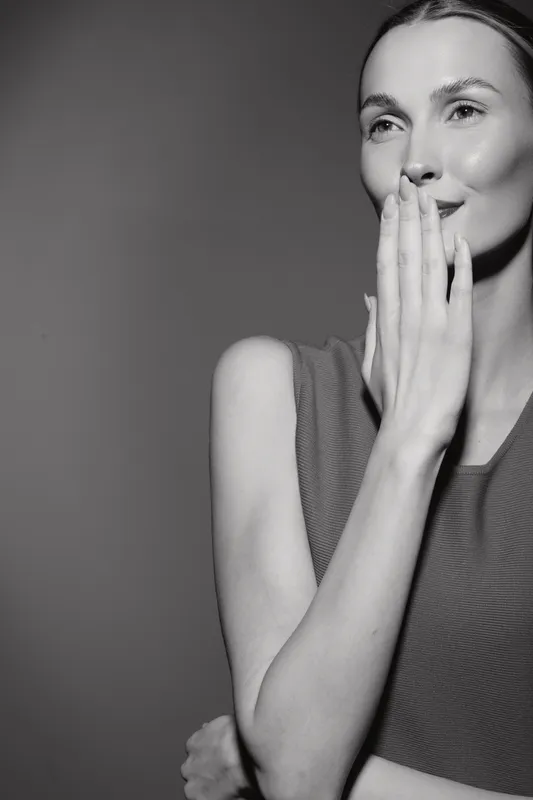 Caucasian woman with light skin and brown hair smiles softly with hand covering mouth, wearing sleeveless top. Concept for emotion, happiness, and candid moment.