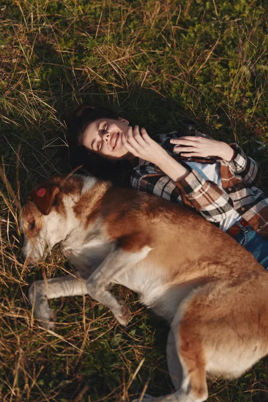 Woman lying on grass beside a large dog in a warm outdoor moment, plaid shirt, relaxed smile, natural light, close companionship, calm nature setting, casual comfort, serene bond.