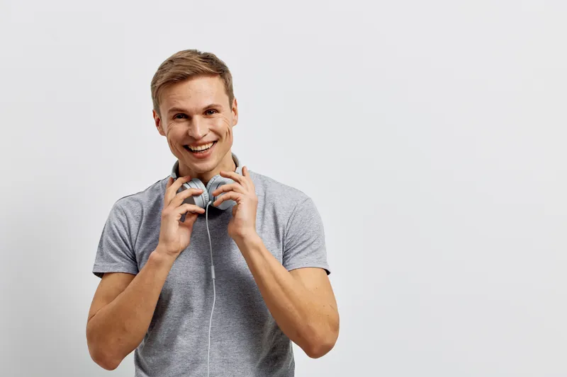 Smiling young man with short hair wearing casual grey t-shirt holds headphones around his neck, looking cheerful, relaxed, and friendly isolated on plain light grey background. People lifestyle concep
