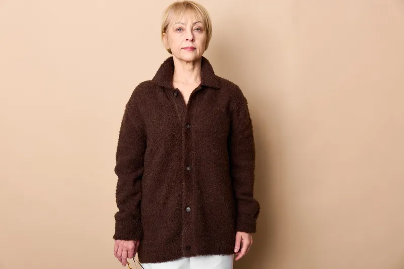 Elderly woman standing in a studio portrait, beige background, wearing a dark cardigan and white pants, calm expression, cozy casual lifestyle, soft light