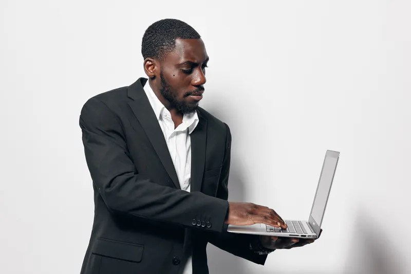 African American man in black suit using laptop computer for work or study on white background. Business professional with dark skin focused on technology and communication.