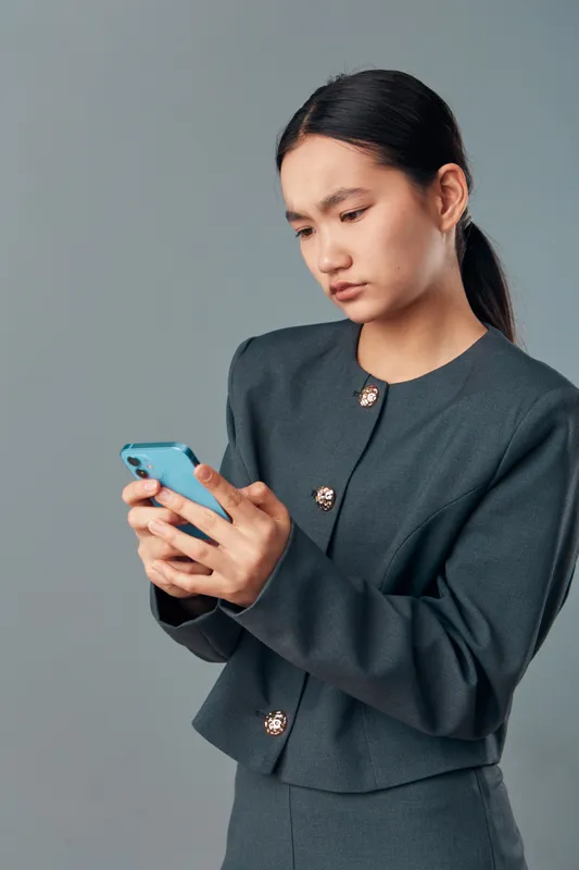 Woman using smartphone with concerned expression, wearing dark formal jacket, standing against gray background. Closeup of young adult focused on mobile device interaction.