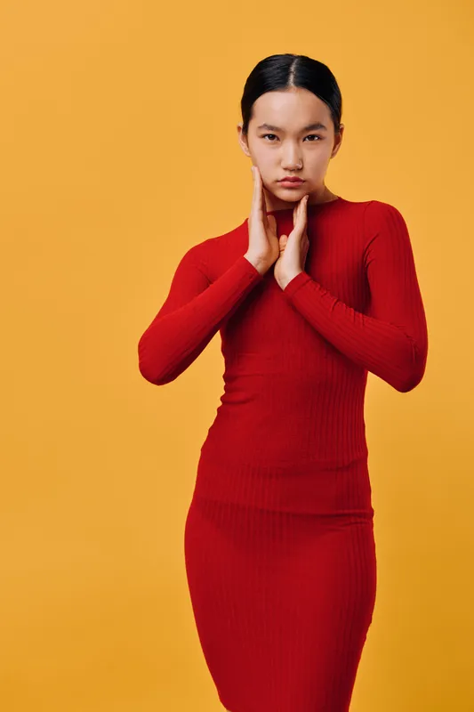 Young woman in red long sleeve dress posing with hands near neck on orange background. Fashion model with serious expression wearing stylish outfit in studio portrait.