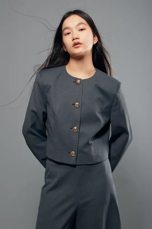 Young woman with long hair wearing gray jacket and pants posing standing with hands behind back in studio. Fashion model with casual outfit looking confident and natural on gray background.