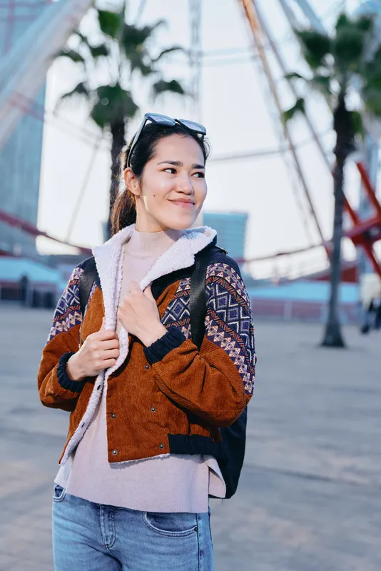 Woman with backpack wearing a warm jacket and sunglasses on head, strolling a sunny urban street, casual fashion and travel vibe, confident smile against a modern city backdrop